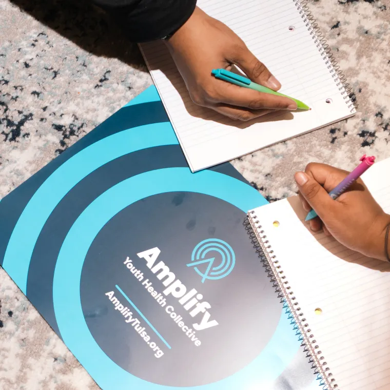 Amplify Youth Health Collective folder on a table beside two people writing in notebooks